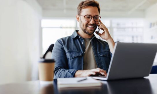 casually-dressed-white-man-with-glasses-smiling-as-he-talks-on-his-smartphone-and-uses-a-laptop